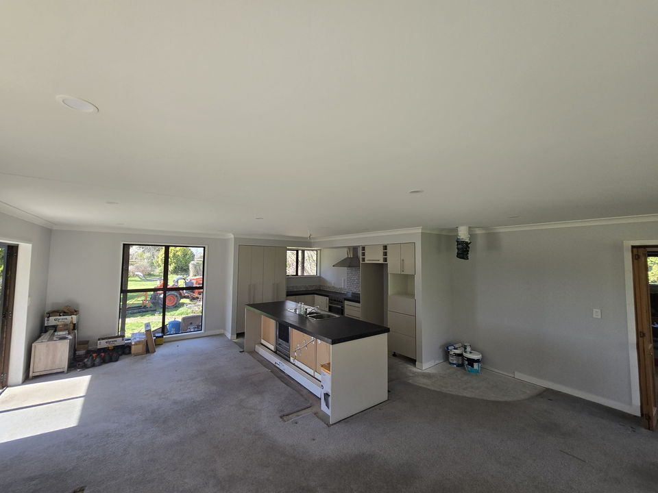 Open-plan living area part way through renovation: walls and ceiling freshly painted, the new kitchen and island fitted with the protective film still on, and big windows onto a lawn